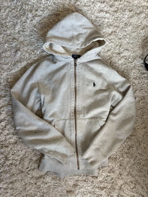 Ljusgrå hoodie från Polo Ralph Lauren - Ljusgrå hoodie med dragkedja från Polo Ralph Lauren. Klassisk design med huva, känguruficka och broderad logga på bröstet. Tillverkad i mjukt bomullsmaterial som är skönt mot huden. Perfekt för en avslappnad och stilren look.