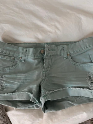 Ljusa gröna jeansshorts med slitningar - Snygga ljusgröna jeansshorts med råa kanter och slitna detaljer framtill. Klassisk femficksmodell med knapp och dragkedja. Perfekta för varma dagar och en avslappnad stil.