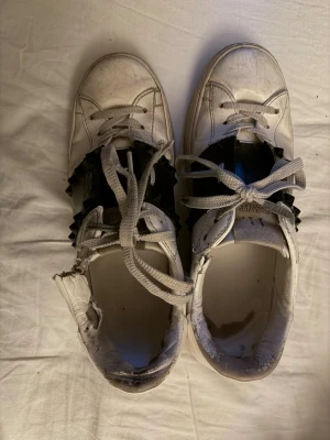 Vita sneakers med svarta detaljer - Snygga vita sneakers från Valentino med bred svart rem över vristen och coola nitar. Skorna har klassisk låg profil, vita snören och är gjorda i läder. Perfekta för dig som gillar streetstyle och vill sticka ut med en unik detalj.
