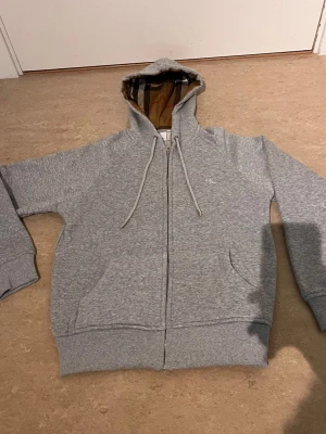 Grå zip-hoodie från Burberry - Snygg grå hoodie med dragkedja från Burberry. Klassisk huva med det ikoniska rutmönstret på insidan, dragsnören och broderad logga på bröstet. Mjuk bomullskänsla och två fickor framtill. Perfekt för en chill och stilren look.