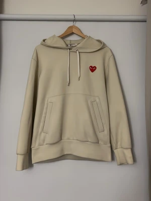 Comme des Garçons hoodie - Pris kan diskuteras! Snygg beige hoodie från Comme des Garçons Play med klassisk huva och dragsko. På bröstet sitter det ikoniska röda hjärtat med ögon. Hoodien har känguruficka och ribbade muddar. Perfekt för en chill och stilren look.