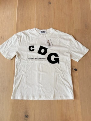 Vit CDG t-shirt från Comme des Garçons - Vit t-shirt från Comme des Garçons Play med stor svart CDG-logga framtill. Klassisk passform med rund hals och korta ärmar. Tillverkad i mjuk bomull, perfekt för en clean och stilren look.