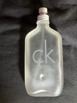 CK One parfymflaska från Calvin Klein -  Parfym från Calvin Klein, modell CK One. Flaskan har en stilren, genomskinlig design med silverfärgad skruvkork och tydlig logotyp på framsidan. Perfekt för dig som gillar klassiska och fräscha dofter.