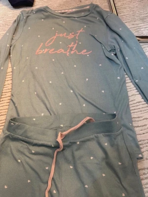 Blå pyjamas med text och hjärtan - Mysig blå pyjamas med långärmad tröja och byxa. Tröjan har texten 'just breathe' i rosa och små vita hjärtan över hela setet. Byxan har rosa dragsko i midjan. Perfekt för chillkvällar hemma.