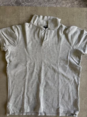 Grå pikétröja med krage - En klassisk grå pikétröja med korta ärmar och krage. Tröjan har knappar framtill och är tillverkad i mjukt bomullsmaterial. Perfekt för en avslappnad och stilren look.