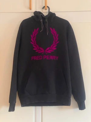 Svart Fred Perry hoodie med lila tryck - Svart hoodie från Fred Perry med stor lila lagerkrans och logga på bröstet. Klassisk modell med huva, dragsnören och känguruficka framtill. Perfekt för en avslappnad och stilren streetlook.