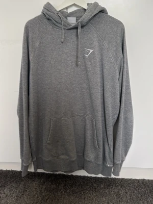 Grå hoodie från Gymshark - Snygg grå hoodie från Gymshark med vit logga på bröstet. Klassisk modell med huva, dragsnören och stor magficka. Perfekt för chill eller träning, i mjukt och bekvämt material. Långa ärmar och ribbade muddar för extra komfort.