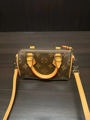 Louis Vuitton Monogram Nano Speedy - Säljer en Louis Vuitton Nano Speedy handväska i klassiskt brunt monogrammönster med beige och gula läderdetaljer. Väskan har guldfärgade metalldetaljer, avtagbar axelrem och två små handtag. Perfekt för dig som gillar lyxiga accessoarer. Köpte den via vinted för 12 tusen. Box och dustbag medföljer!💕