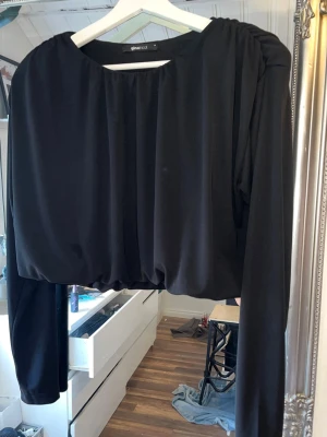 Svart croppad topp från Gina Tricot - Stilren svart croppad topp från Gina Tricot med långa ärmar och rund halsringning. Toppen har en lös passform och är tillverkad i ett mjukt, lätt material som ger ett snyggt fall. Perfekt att matcha med höga jeans eller kjol.