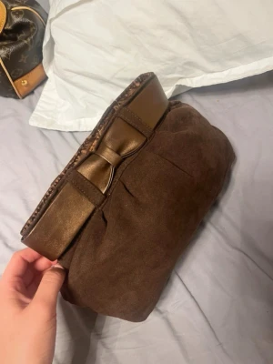 Brun kuvertväska med rosett - Snygg brun kuvertväska i mockaliknande material med metallicbrun kant och en dekorativ rosett framtill. Väskan har en mjuk, puffig form och stängs upptill. Perfekt accessoar för att lyfta din outfit. Brun clutch väska med rosett i mocka imitation. Det står inget märke på den men den är köpt på en marknad!