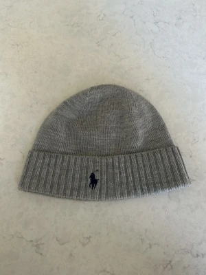 Grå mössa från Polo Ralph Lauren - Snygg grå ribbstickad mössa från Polo Ralph Lauren i ull. Klassisk design med uppvikt kant och broderad logga framtill. Perfekt accessoar för att hålla värmen och ge en stilren touch till din outfit.