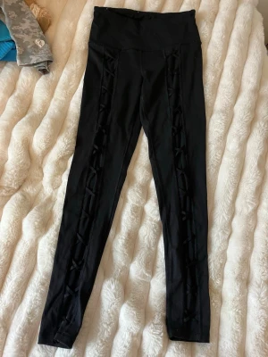 Svarta leggings med snörningsdetaljer - Snygga svarta leggings med hög midja och dekorativa snörningsdetaljer längs benen. Tight passform och stretchigt material som sitter bekvämt. Perfekta för dig som vill ha en stilren men ändå unik look. 