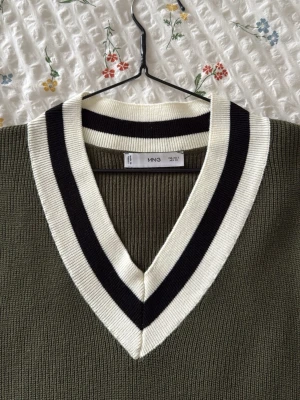 Olivgrön stickad väst från MNG -  olivgrön stickad väst från MNG med bred v-ringning. Västen har vita och svarta ränder runt halsen och ärmarna för en sportig look. Perfekt att styla över en skjorta eller t-shirt för en trendig outfit.