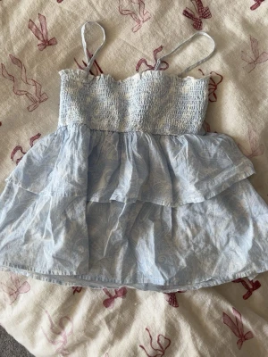 Ljusblå volang jumpsuit med smock - Supersöt ljusblå jumpsuit med smala axelband, smockad överdel och dubbla volanger. Shortsdelen är luftig och tyget har ett diskret mönster. Perfekt för sommardagar och ger en riktigt fräsch look. Materialet känns som bomull eller linne.