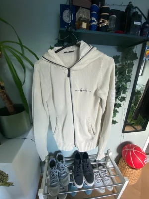  Sail Racing - Snygg vit hoodie från Sail Racing med dragkedja framtill och två fickor. Tillverkad i ett stickat material med mesh på insidan och har en stor huva. Perfekt för en avslappnad och sportig stil.