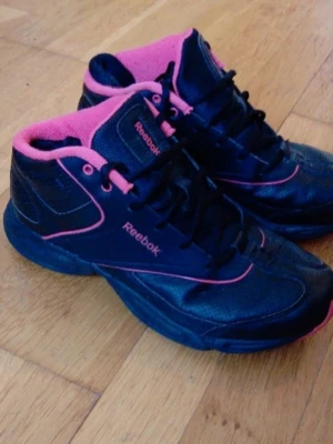 Svarta och rosa Reebok sneakers - Snygga svarta sneakers från Reebok med rosa detaljer och foder. Skorna har en klassisk mid cut-design, svarta skosnören och Reebok-logga på sidan och plösen. Perfekta för dig som gillar sportig stil och vill sticka ut med färg.