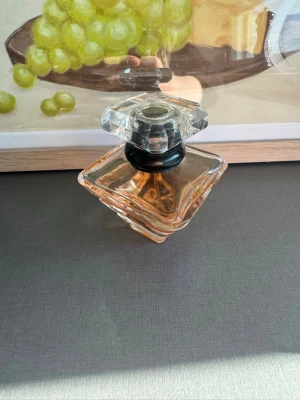 Elegant parfymflaska i glas - Säljer en elegant parfymflaska i transparent glas med fyrkantig form och tjock, kristalliknande kork. Flaskan har en svagt rosa-orange vätska inuti och en svart detalj vid halsen. Perfekt för dig som gillar snygga och stilrena parfymer.