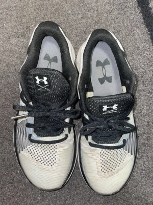 Under Armour svarta och vita sneakers - Snygga sneakers från Under Armour i svart och vitt med meshdetaljer och klassisk logga på plösen och sidan. Skorna har lågt skaft, svarta snören och en sportig design med ventilerande material och bekväm sula. Perfekta för en aktiv livsstil.