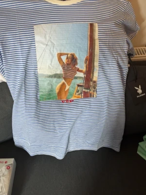 Blåvit randig t-shirt från Playboy OBS Exklusiv utgåva  - Snygg blåvit randig t-shirt från Playboy med tryck av en retroinspirerad bild på framsidan. T-shirten har rund halsringning och korta ärmar. Perfekt för dig som gillar streetstyle och vill sticka ut lite extra.