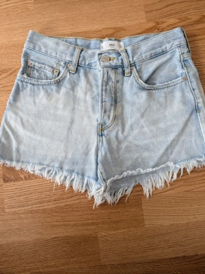 Ljusblå denimshorts från MNG - Säljer ett par ljusblå denimshorts från MNG med fransig kant och klassisk femficksdesign. Shortsen har hög midja och är perfekta för sommaren. Snyggt slitna detaljer och en avslappnad passform.