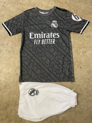 Real Madrid Bellingham matchställ - Snyggt matchställ från Real Madrid med Bellingham och nummer 5 på ryggen. Tröjan är grå med svart mönster, vita detaljer och klubbmärke samt sponsortryck. Shortsen är vita med klubbmärke. Perfekt för fotbollsträning eller match.