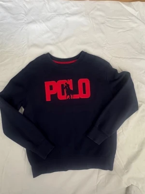 Mörkblå sweatshirt från Polo Ralph Lauren - Snygg mörkblå sweatshirt från Polo Ralph Lauren med stor röd POLO-logga och ikonisk ryttare på bröstet. Tröjan har rund halsringning, ribbade muddar och är tillverkad i mjukt material. Perfekt för en avslappnad och stilren look.