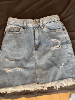 Blå jeanskjol med slitningar - Trendig blå jeanskjol i kort modell med slitna detaljer och fransad nederkant. Klassisk femficksdesign och bälteshällor. Perfekt för dig som gillar en avslappnad och cool stil.