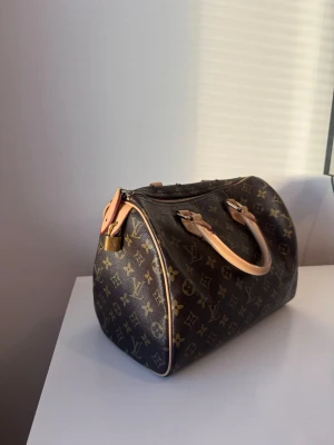 Louis Vuitton Speedy Monogram väska - Klassisk Louis Vuitton Speedy handväska i brunt monogramcanvas med beige läderhandtag och guldfärgade metalldetaljer. Ikoniskt LV-mönster över hela väskan och dragkedja upptill. Perfekt för dig som gillar lyxiga accessoarer. Nyckellås tillkommer inte.