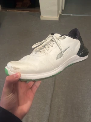 Vita golfskor från Puma med dubbar - Säljer ett par vita golfskor från Puma med gröna sulor och svarta detaljer. Skorna har snörning, slitstarka dubbar undertill och märkningen 'NITRO' på hälen. Perfekta för golfbanan med bra grepp och sportig look. Nästan helt nya, endast använda en halv golf sommar, nypris 1999kr