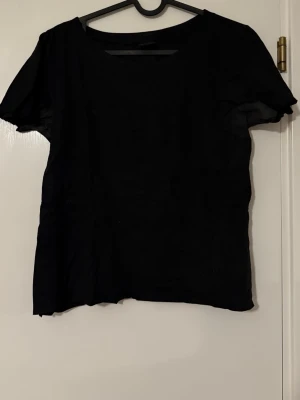 Svart t-shirt från Gina Tricot i suede/mocka - En klassisk svart t-shirt från Gina Tricot i enkel och stilren design. T-shirten har rund halsringning och korta ärmar, perfekt att matcha med jeans eller kjol. Tillverkad i mjuk bomull för en bekväm känsla.