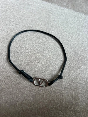 Svart armband från Valentino - Stilrent svart armband från Valentino med justerbart band i textil och en silverfärgad metallberlock med det ikoniska V-logotypen. Perfekt accessoar för att lyfta din outfit med en touch av lyx och enkelhet.