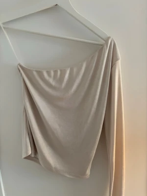 Beige oneshoulder topp - Trendig beige oneshoulder topp med lång ärm. Tunn och mjuk i materialet, perfekt för dig som gillar minimalistisk stil. Sitter snyggt och ger en cool asymmetrisk look.
