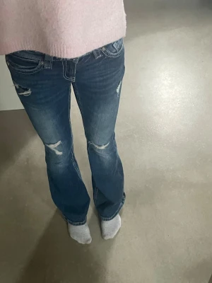 Blå bootcut jeans med slitningar - Snygga blå bootcut jeans med slitningar på knän och lår. Jeansen har låg midja, klassiska fem fickor och en lätt tvättad look. Perfekta för en avslappnad och trendig stil.