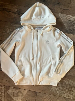Vit hoodie från Adidas med dragkedja - Snygg vit hoodie från Adidas med dragkedja framtill och klassiska tre ränder i silver längs ärmarna. Tröjan har huva med dragsko, två fickor med knappar och ribbade muddar. Perfekt för en sportig och avslappnad stil.