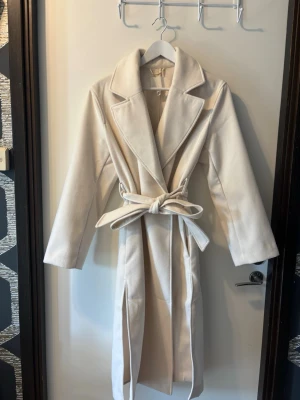 Beige lång kappa med knytskärp - Stilren beige kappa med lång passform och breda slag. Kappan har ett matchande knytskärp i midjan och raka ärmar. Perfekt för dig som gillar minimalistisk och klassisk stil. Materialet ser ut att vara mjukt och ger en lyxig känsla.