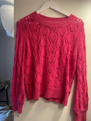 Rosa stickad tröja med hålmönster - Säljer en rosa stickad tröja med snyggt hålmönster och bred ribbad mudd vid hals, ärmslut och nederkant. Tröjan har lång ärm och en avslappnad passform. Perfekt för dig som vill ha något både trendigt och bekvämt.
