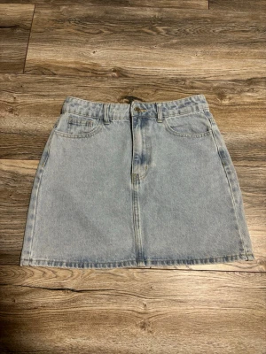 Ljusblå jeanskjol i klassisk modell - Snygg ljusblå jeanskjol med klassisk femficksdesign och bälteshällor. Kjolen har en rak passform och stängs med dragkedja och knapp framtill. Perfekt att matcha med en t-shirt eller hoodie för en avslappnad look.