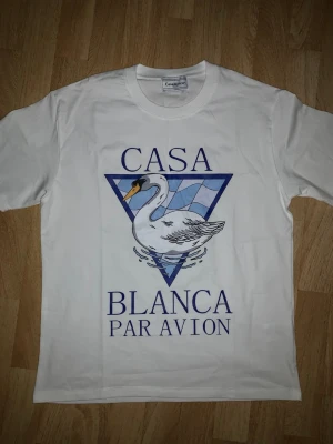 Vit Casablanca t-shirt med svanmotiv - Vit t-shirt från Casablanca med tryck av en svan och texten 'Casa Blanca Par Avion' på framsidan. Klassisk rund hals och korta ärmar. Tillverkad i mjuk bomull, perfekt för en avslappnad och stilren look.