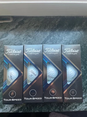 Titleist Tour Speed golfbollar - Fyra förpackningar Titleist Tour Speed golfbollar. Bollarna är vita med klassisk dimple-yta och har Titleist-loggan tryckt på sig. Förpackningarna är svarta med blå detaljer och fönster som visar bollen inuti. Perfekt för dig som vill ha kvalitetsbollar till golfbanan. Bollarna är helt nya och fina plus att du får med en greenlagare. 