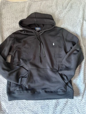 Svart hoodie från Polo Ralph Lauren - Svart hoodie från Polo Ralph Lauren med klassisk huva och dragsko. Har en stor ficka framtill och den ikoniska broderade loggan i vitt på bröstet. Mjuk bomullskänsla och stilren design som passar perfekt till jeans eller joggers.