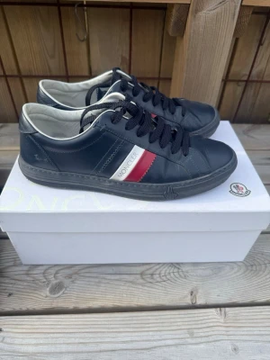 blåa Moncler sneakers  - Snygga marinblå sneakers från Moncler i läder med klassiska ränder i rött och vitt på sidan samt Moncler-logga på plösen. Skorna har svarta snören och vit insida, perfekt för dig som gillar stilrena och exklusiva sneakers.