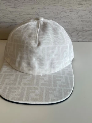 Vit keps från Fendi med FF-mönster - Snygg vit keps från Fendi med det klassiska FF-monogrammet ton-i-ton över hela kepsen. Kepsen har böjd skärm och är tillverkad i ett tygmaterial som känns både lyxigt och stilrent. Perfekt accessoar för att lyfta din outfit.