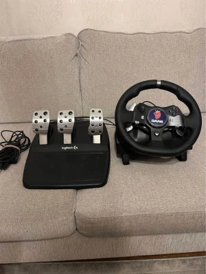 Logitech ratt och pedaler med Saab-emblem - Säljer en svart Logitech ratt med Saab-emblem och tillhörande pedalset i metall och plast. Ratten har flera knappar och är klädd i konstläder, perfekt för racing på konsol eller PC. Pedalerna har tre metallpedaler för realistisk körkänsla. Levereras ej, finns att hämta i falköping 