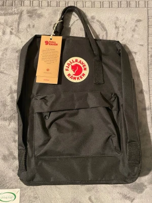 Svart Kånken ryggsäck från Fjällräven - Klassisk svart Kånken ryggsäck från Fjällräven med rund röd logotyp framtill. Ryggsäcken har ett stort huvudfack, en frontficka med dragkedja och justerbara axelband. Tillverkad i slitstarkt syntetmaterial och har topphandtag för enkel bärning.