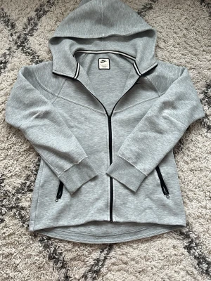 Grå zip-hoodie från Nike - Säljer en grå hoodie från Nike med hel dragkedja och svarta detaljer. Hoodien har två fickor med dragkedja, ribbade muddar och Nike-logga på bröstet. Materialet är mjukt och bekvämt, perfekt för chill eller träning. Snygg kontrast med svart dragkedja och kantband vid kragen.