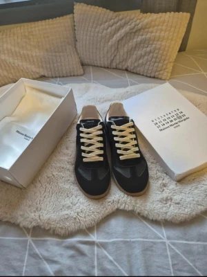 Maison Margiela svart sneaker - Maison Margiela sneakers i svart läder med beige detaljer och vita snören. Klassisk låg modell med gummisula och stilren design. Märkets ikoniska logotyp på tungan och insidan. Perfekt för dig som gillar exklusiva och trendiga skor.