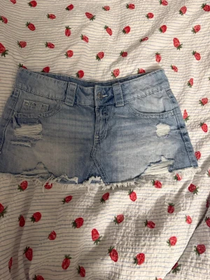 Blå jeanskjol med slitningar - Cool kort jeanskjol i ljusblå denim med slitna detaljer och rå kant. Kjolen har klassiska fickor fram och bak samt bälteshällor. Perfekt för dig som gillar en avslappnad och trendig look.