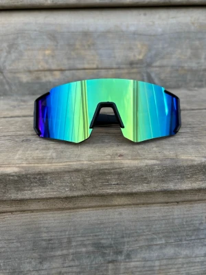 Barn grön /blå Sportiga solglasögon med spegelglas - Barnstorlek, kan även passa äldre med litet ansikte. Säljer ett par sportiga solglasögon med stora, spegelblå och gröna linser. De har en futuristisk och kantig form med svart ram. Perfekta för dig som vill ha en cool look och skydda ögonen mot solen.