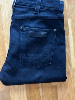 Svarta jeans från Wrangler - Säljer ett par svarta jeans från Wrangler med klassisk femficksdesign och snygga detaljer på bakfickan. Jeansen har raka ben och är tillverkade i slitstark denim. Perfekt för dig som gillar en tidlös och cool look.