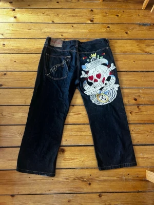 Baggy Ed hardy-jeans med stort broderi på baksidan  - Baggy jeans från Ed Hardy med stor broderad dödskalle på baksidan. Raka ben, rätt så lågmidjade och klassisk femficksmodell. Materialet är kraftig denim och byxorna har coola detaljer som broderad signatur på bakfickan. Lite till inget slitage, se bilder (lite slitage nere påvänstra benet och en maska dragen precis under broderiet). Köpta för 2500 kr säljes för 1500 kr 
